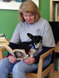 An older woman sits in a chair, reading a book with a small black and white dog on her lap. The woman is wearing a gray t-shirt and jeans. Bookshelves and a green wall are in the background.