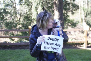A woman in a black jacket holds a small dog licking her face. She smiles and holds a sign that reads Doggy Kisses Are The Best! They are outdoors with trees and a wooden fence in the background.