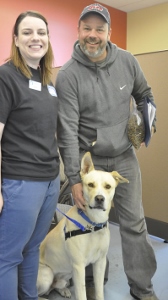 Two people smile at the camera while standing next to a large yellow dog wearing a harness. The man is petting the dog, and the background features colorful walls.