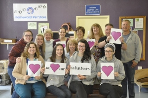 A group of smiling people, some seated and some standing, hold signs with pink hearts and the words We ♥ our volunteers. They are indoors in front of a sign reading Homewood Pet Store.