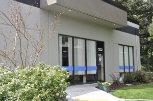 A modern building with large windows featuring blue horizontal stripes, surrounded by greenery and shrubs, with a small paved walkway leading to the entrance.