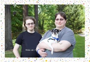 Two people stand outside surrounded by trees; one holds a small black and white dog. Both are smiling and wear glasses. The image has a white border with colorful confetti dots.