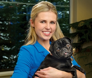 A smiling woman with long blonde hair, wearing a blue top, holds a small black pug in her arms indoors with a green plant and window in the background.