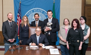 A group of people stands behind a seated man signing a document at a desk, with U.S. and state flags and an official seal in the background. One person holds a small dog.