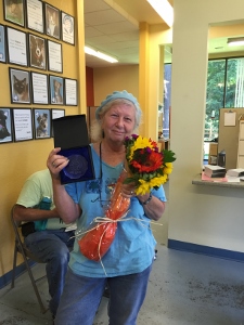 An older woman wearing a blue shirt and hair net holds a bouquet of flowers and a boxed award, smiling in a brightly lit room with animal photos on the wall behind her.