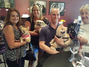 Four smiling adults, three women and one man, each holding small dogs, pose together indoors. Two empty wine glasses sit on the table in the foreground. Art hangs on the red wall behind them.