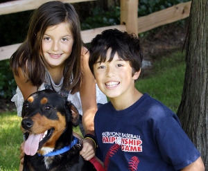 Two smiling children, a girl and a boy, kneel on grass next to a happy black and brown dog wearing a blue collar. A wooden fence and trees are visible in the background.