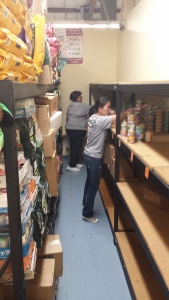 Two people organize canned goods and other food items on shelves in a storage room, with boxes and bags stacked on shelves along the opposite wall.