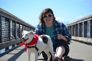 A person wearing sunglasses and a plaid shirt sits on a boardwalk, smiling next to a white dog with a red bandana around its neck on a sunny day.
