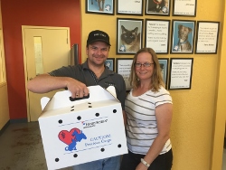A man and a woman smile while holding a white pet carrier box with a red heart and blue cat logo. Framed cat photos and descriptions are displayed on the yellow wall behind them.