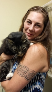 A woman with long brown hair and a patterned tattoo on her upper arm holds a large, fluffy black and white cat. She is wearing sunglasses on her head and a blue and white sleeveless top.