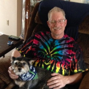 A smiling man wearing glasses and a colorful tie-dye shirt sits in a brown recliner, holding a small black and gray dog with a patterned blue bandana on its neck.