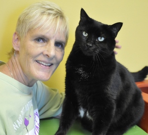 A smiling person with short blond hair is sitting next to a large black cat. The person is wearing a light green shirt, and the background is yellow. The cat has green eyes and is looking at the camera.