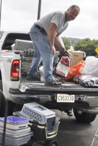 A man stands on the bed of a pickup truck, unloading various pet supplies and bags. Several pet carriers are on a cart next to the truck, and the man is holding a bag of pet food.