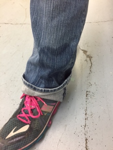 A person wearing blue jeans and a dark sneaker with pink laces stands on a light-colored floor with some dirt or scuff marks. The jeans are slightly cuffed at the bottom.