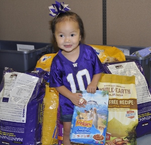 A young girl wearing a purple sports jersey and matching bow stands in front of large bags of pet food, smiling and holding a bag of Friskies cat treats.