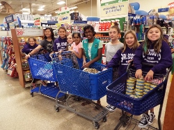 A group of children and an adult stand in a store aisle with three blue shopping carts filled with pet supplies, smiling at the camera. Shelves of pet products are visible in the background.