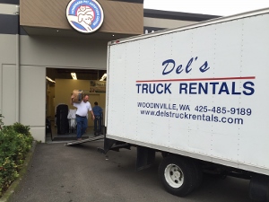 A moving truck labeled Dels Truck Rentals is parked outside a building. Two men are carrying a large item from inside the building towards the truck. The business sign is visible above the entrance.