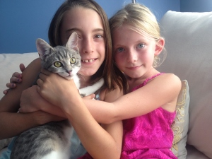Two young girls sitting on a couch, both smiling and holding a gray and white cat. One girl wears a pink dress, while the other wears a light blue top. They are in a cozy, brightly lit room.