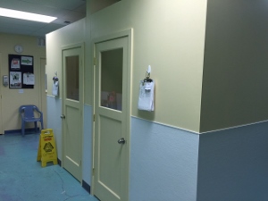 Two closed doors with clipboards attached are seen in a hallway with pale yellow and blue walls. A gray chair, a bulletin board, and a yellow caution sign are visible in the background. The area appears to be a waiting or clinic room.