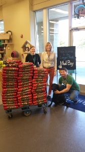 Three people pose inside a pet store with a cart stacked high with bags of pet food. One person crouches to pet a black dog. There is a welcome sign in the background that says Welcome Cougar Pack Mud Bay.