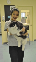 A smiling person stands indoors holding a large, fluffy black and white cat in their arms. There are doors, a clock, and chairs visible in the background.