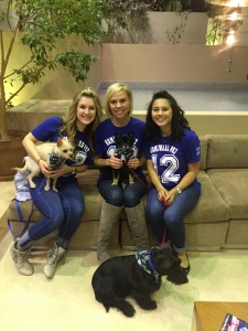 Three women sit on a couch, each wearing a blue t-shirt and holding small dogs. Another black dog sits in front of them on the floor with a red leash. There are plants and a beige sofa in the background.