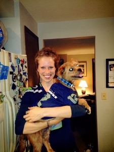 A smiling woman wearing a Seattle Seahawks jersey holds a brown dog in her arms inside a cozy kitchen. The dog wears a blue collar and looks off to the side. Refrigerator magnets are visible in the background.