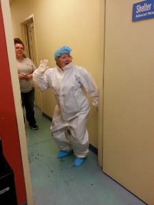 A person in a white protective suit, blue hair cover, and shoe covers poses playfully in a hallway near a door labeled Shelter. Another person stands in the background, smiling.