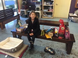 A young girl sits on a wooden bench in a room with pet supplies, including food, toys, and a dog bed, arranged around her. Animal crates are stacked in the background.
