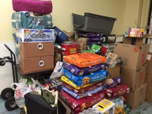 A stack of pet food bags, toilet paper, cardboard boxes, and plastic bags piled against a wall, with a dolly cart nearby in a storage room.