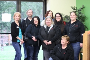A group of eight adults, most wearing dark jackets, pose together indoors near a window and green wall with plants. Some are standing and others are kneeling, all smiling at the camera.