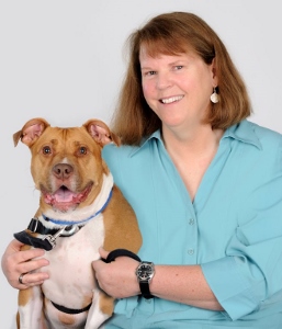 A smiling woman with shoulder-length brown hair, wearing a light blue shirt, poses with a happy brown and white dog wearing a harness and collar.