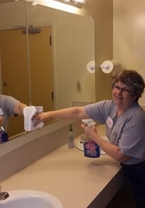 A person wearing glasses and a gray shirt is smiling while cleaning a bathroom mirror with a cloth and a bottle of glass cleaner.