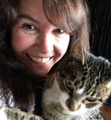 A woman with long brown hair smiles at the camera while holding a tabby cat with green eyes in her arms.