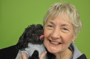 An older woman smiles while holding a small black dog in a gray sweater. The dog is licking her cheek. The background is solid green.