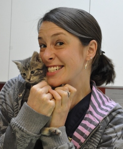A woman with dark hair smiles widely while holding a small gray tabby kitten close to her face, both appearing happy and content.