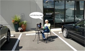 A woman sits at a small outdoor table with a dog in a parking lot. The dog has a speech bubble saying, Isnt this fun, Bob? There are parked cars, a plant, and a building in the background.