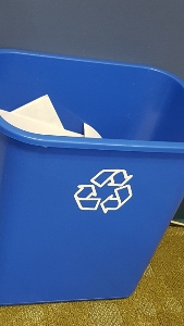 A blue recycling bin with the white recycling symbol on the side, containing a few sheets of white paper, sits on a carpeted floor against a blue wall.