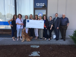 A group of ten people and a golden retriever pose outside a building, holding a large ceremonial check. The building number 13132 is visible above the door.