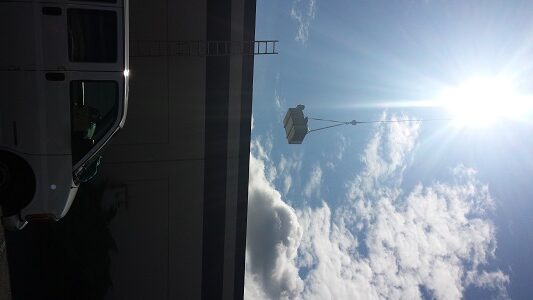 A white van is parked next to a building while an HVAC unit is lifted by a crane in the bright, sunny sky with scattered clouds. A ladder leans against the buildings roof.