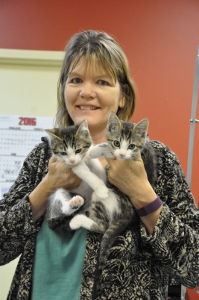 A woman smiling while holding two gray and white kittens in her arms, standing in front of a red wall and a calendar.
