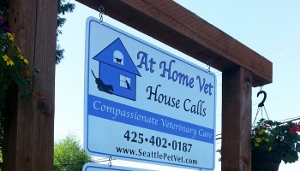 A sign for At Home Vet House Calls featuring animal silhouettes, the text Compassionate Veterinary Care, a phone number, and a website address. The sign is attached to a wooden post outdoors.
