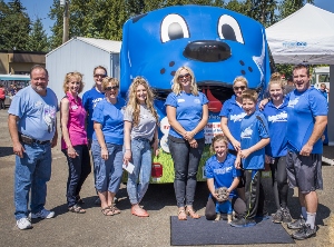 A group of people pose and smile in front of a large blue cartoon dog vehicle at an outdoor event on a sunny day. Most are wearing blue shirts and there are trees and tents in the background.