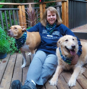 A woman sits on a wooden deck with a brown-and-white dog and a golden retriever, both wearing matching patterned bandanas. She is smiling and wearing a blue Seahawks Champions sweatshirt.