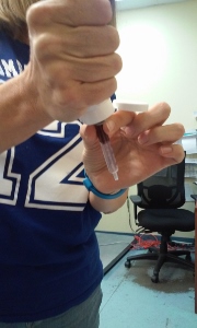 A person wearing a blue shirt with the number 14 is using a pipette to transfer a red liquid into a small tube in a laboratory setting. An office chair and desk are visible in the background.
