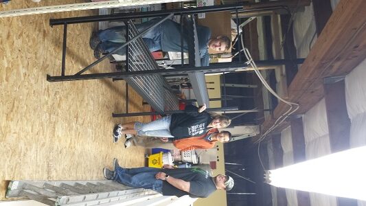 Four people are standing and sitting on a black metal structure inside a workshop with wooden floors and exposed beams. Tools and equipment are visible in the background.