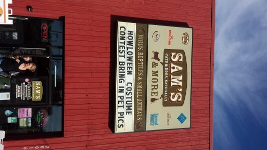A red building with a sign reading “Sam’s Pets & More” and smaller text about birds, reptiles, small animals, and a Halloween costume contest for pets. The store entrance and an open sign are visible on the left.