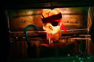 A skeleton with a red bandana and eye patch, holding a hook, is lit with eerie orange and green lights inside a wooden chest, creating a spooky, pirate-themed scene.