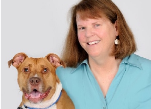 A smiling woman with shoulder-length brown hair in a light blue shirt sits next to a happy brown dog with a white chest and blue collar, both facing the camera against a plain light background.